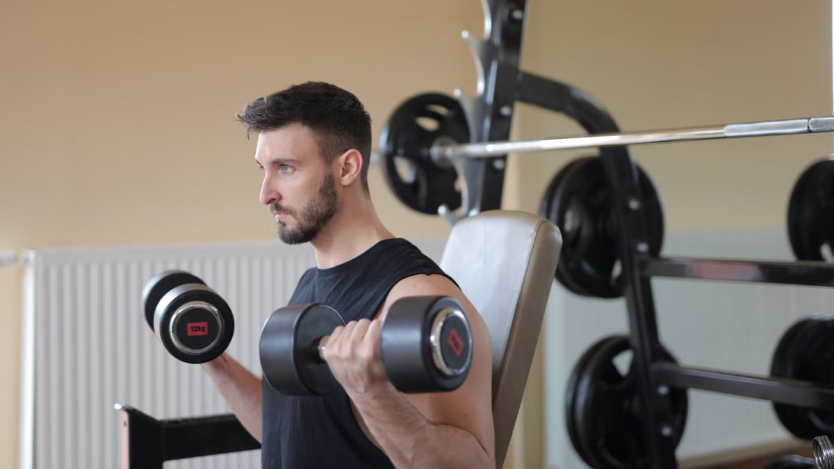 Focused man lifting dumbbells for bicep curls in gym, emphasizing strength and fitness.